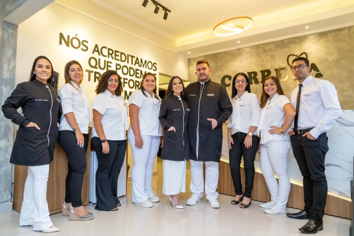 Oito integrantes da equipe da Correia Odontologia, de jaleco e uniforme, posando na recepção diante da parede com a frase "Nós acreditamos que podemos transformar seu mundo"