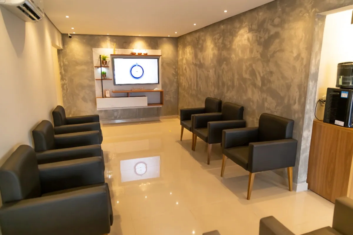Sala de espera com poltronas escuras enfileiradas e televisão na parede